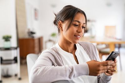 Mature african woman sitting on couch at home using smartphone. Black woman relaxing on the sofa while messaging with a cellphone at home. Happy mature indian woman typing on mobile phone in living room.