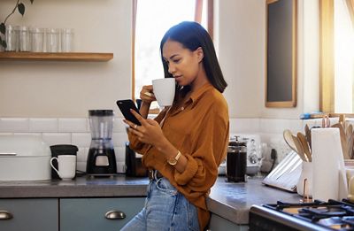 Young mixed race woman drinking a cup of coffee while using her phone alone in the morning in the kitchen. Happy hispanic female smiling and enjoying a cup of tea while using social media on her phone at home