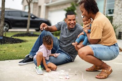 An African American family enjoys a sunny day outdoors drawing with chalk. Parents are interacting with their children, smiling and bonding. The scene exudes joy, love, and the essence of family togetherness.