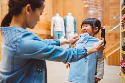 Pretty young mom fitting a shirt on the cheerful little girl at the kid's wear department in a fashion boutique