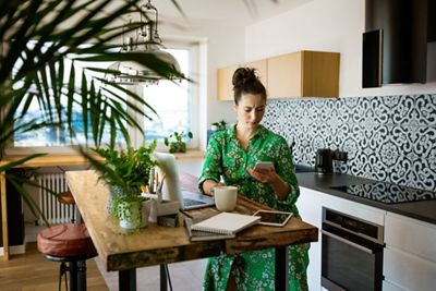 Pregnant young beautiful woman wearing green dress sitting at table in the kitchen and working at home, using smart phone.
