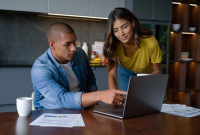Couple paying bills online from home on a laptop computer and man pointing at the screen looking concerned about the expenses. **DESIGN ON DOCUMENTS BELONG TO US**