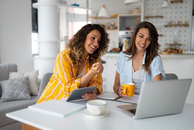 Joyful sisters using digital tablet and laptop for work from home while drinking tea