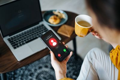 Over the shoulder view of young Asian woman receiving an incoming suspected call from unknown caller on her smartphone and rejecting the call at home. Device screen showing warning sign as detected by the network provider. Phone scam and fraud concept