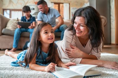 A mother and daughter are lying on their tummies on the floor reading a book together, while dad and their son are watching a digital screen in the background