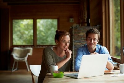 Smiling young couple using laptop while having coffee at table in cottage